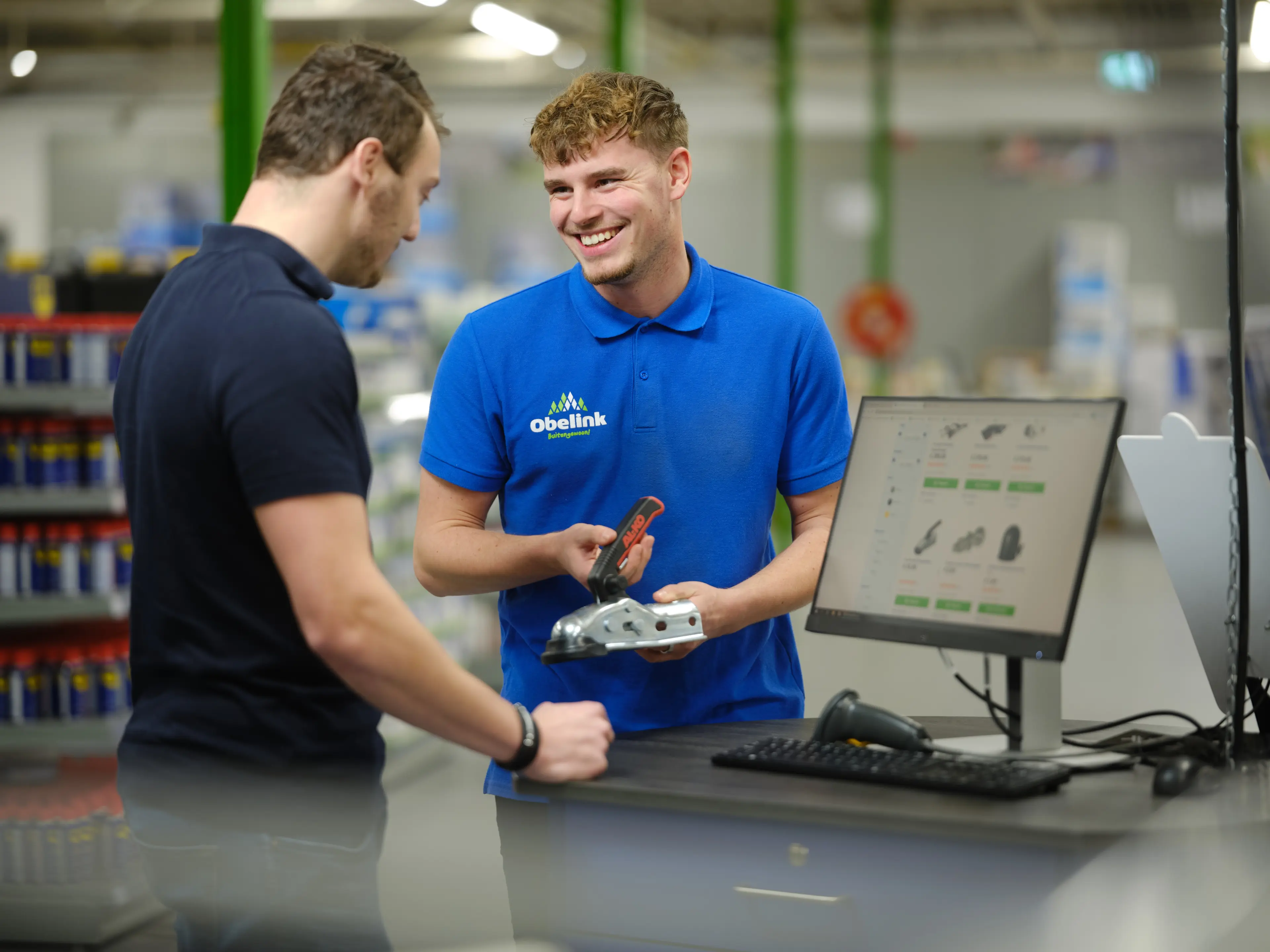 Kom je er niet helemaal uit? Medewerker in blauw Obelink poloshirt toont een metalen bevestigingsonderdeel aan klant bij verkoopbalie met computerscherm waarop producten zichtbaar zijn.